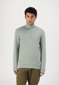 Sudadera verde claro de cuello alto hecha de una tela suave; presenta un cierre de cremallera de un cuarto en la parte delantera y puños acanalados, combinada con pantalones marrones sueltos.