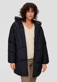 Vrouw met krullend haar, gekleed in een zwarte pufferjack over een beige V-hals trui en geruite broek, staand tegen een effen grijze achtergrond.