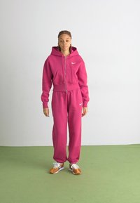 Fuksijas rozā kapucēta svītra ar priekšējo kabatu un atbilstošām sviedru biksēm. Balts Nike logo uz krūšu daļas. Modelis valkā brūnas un baltas sporta apavus.