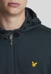 Marineblauer Reißverschluss-Hoodie mit einer Kordelzugkapuze und einem kleinen gelben Vogel-Logo auf der Brust, hergestellt aus einem strukturierten, atmungsaktiven Material.