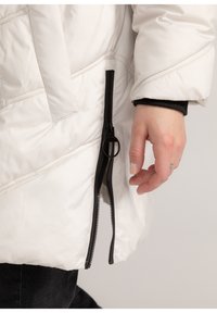 Witte pufferjas met een diagonaal gestikt patroon, zwarte rits aan de zijkant en een zachte, gladde stoftextuur. Arm zichtbaar aan de rand.