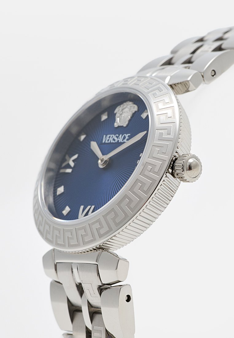 Montre en métal argenté avec un cadran texturé bleu, arborant un logo de lion, un motif de clé grecque sur la lunette et des index horaires en argent.