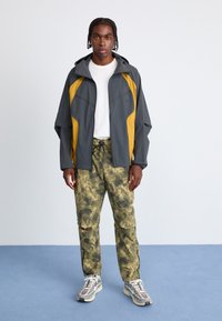 Grå og gul vindjakke med hætte, parret med grønne camo cargo bukser. Grå sneakers med strukturelle detaljer. Rent, minimalistisk design.