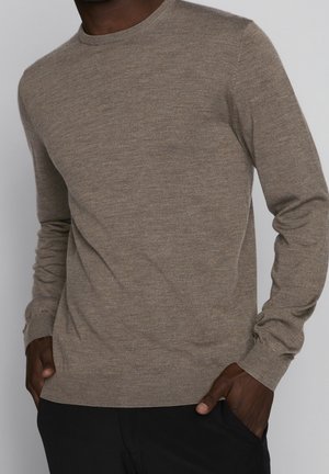 Mann trägt einen enganliegenden beigen Langarmpullover mit den Händen in den Taschen einer schwarzen Hose vor einfarbigem Hintergrund.