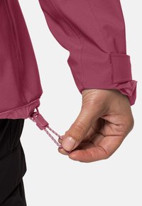 Mano che regola il cordino dell'orlo di una giacca rosa, mostrando la texture del tessuto e il fermo elastico su uno sfondo chiaro.