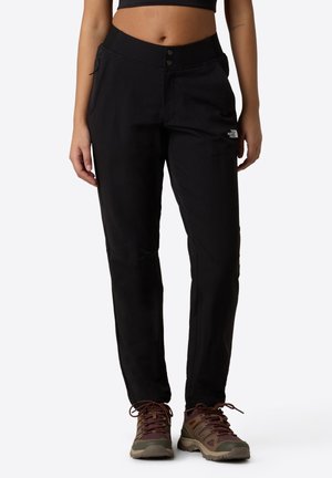 Woman wearing black The North Face hiking pants and brown trail shoes, standing against a plain light background.