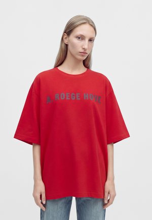 Une femme porte un T-shirt rouge oversize à inscription foncée et un jean bleu sur un fond uni et clair.