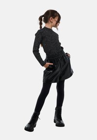 Haut noir à manches longues avec un subtil effet pailleté, associé à une jupe noire brillante avec une ceinture élastique et des poches latérales, porté avec des bottes noires.