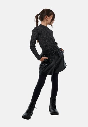 Haut noir à manches longues avec un subtil effet pailleté, associé à une jupe noire brillante avec une ceinture élastique et des poches latérales, porté avec des bottes noires.