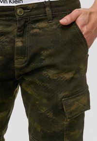 Spodnie cargo w kamuflażu, charakteryzujące się zielono-brązową kolorystyką, kieszeniami z materiału oraz zapięciem na guziki w pasie.