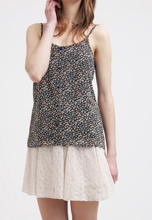 Woman wearing a black floral sleeveless top and a white eyelet skirt, standing with one hand near her face against a plain background.