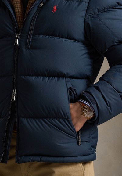 Mand iført navyblå dunjakke med rødt logo, hånd i lommen og synligt brunt armbåndsur med blå- og hvidstribet skjortemanchet.