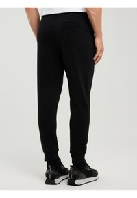 Mees, kes kannab musti kitsenevaid mangurpükse ja musti tenniseid, seisab parema käega külje ääres lõdvestunult, ühtlasel heledal taustal.