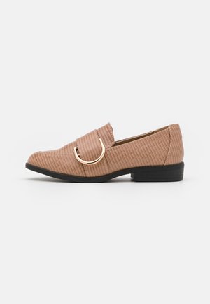 Mocasín beige con textura, tacón bajo negro y detalle de hebilla curva dorada en la parte superior, visto de lado sobre fondo blanco.