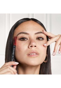 Crayon eyeliner noir avec une pointe rouge, tenu près du visage d'un modèle. Elle a de longs cheveux foncés et porte de grandes boucles d'oreilles rondes. Des taches de rousseur sont visibles.