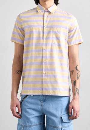 Mann trägt kurzärmeliges, geknöpftes Hemd mit gelben und hellvioletten horizontalen Streifen und hellblaue Jeansshorts mit Taschen.