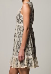 Verkkomekko vaaleanbeigellä, jossa on kukkakuviot, olkaimeton malli, istuva vyötärö ja röyhelöhelma. Teksturoitu, kerroksellinen yksityiskohta helmassa.