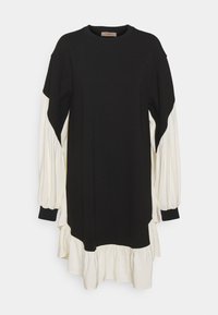 Sort og cremefarvet kjole med rund halsudskæring, lange ballonærmer og en lagdelt flæsekant. Fremstillet af en glat stof med kontrasterende farver.