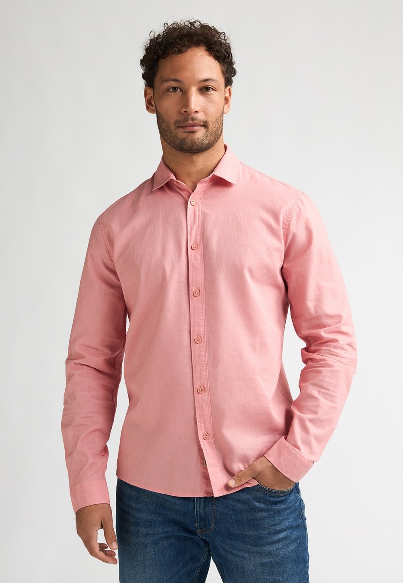 Homme aux cheveux bouclés portant une chemise rose à manches longues boutonnée et un jean bleu, debout avec une main dans la poche devant un fond uni.