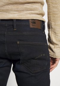 Detailní záběr osoby v tmavě modrých džínách s viditelným zadním kapsou a koženou značkovou nášivkou, doplněné béžovým pleteným svetrem.