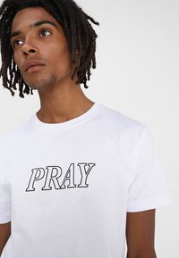 Balta medvilninė marškinė su ryškiu juodu užrašu "PRAY", apvaliu kaklu, trumpomis rankovėmis ir klasikinio silueto. Sklandus paviršius be raštų.