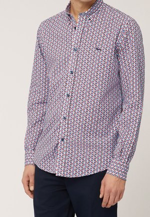 Homme portant une chemise à manches longues boutonnée avec un motif géométrique en losanges rouges, blancs et bleus et un pantalon foncé.