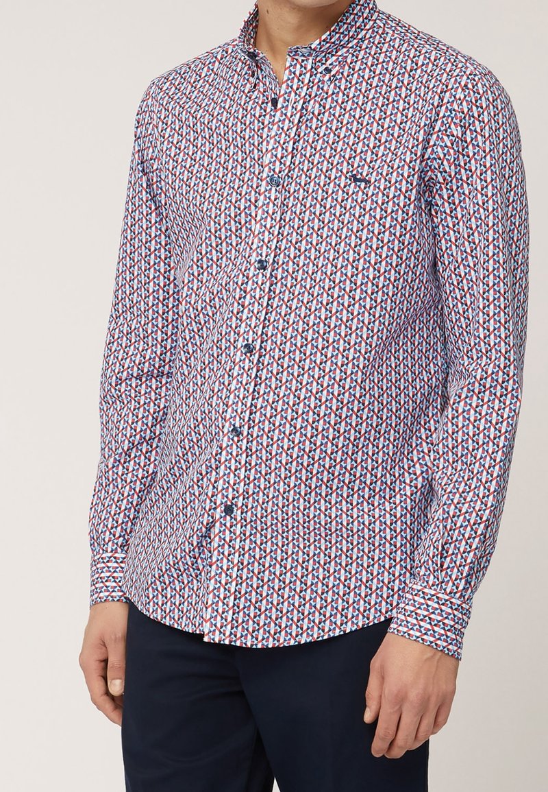 Homme portant une chemise à manches longues boutonnée avec un motif géométrique en losanges rouges, blancs et bleus et un pantalon foncé.