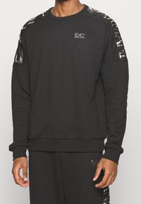 Uomo con felpa a maniche lunghe nera e pantaloni con loghi lucidi Emporio Armani EA7 sul petto, sulle spalle e sulla coscia.