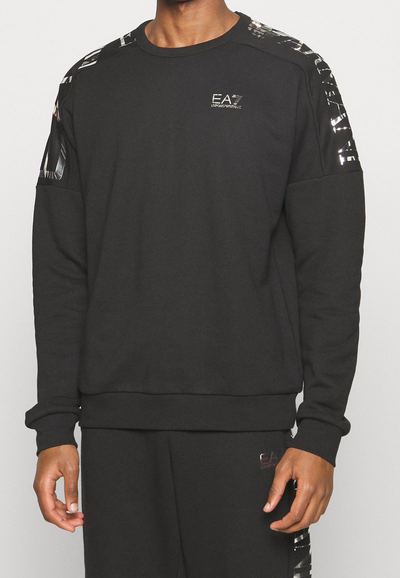 Uomo con felpa a maniche lunghe nera e pantaloni con loghi lucidi Emporio Armani EA7 sul petto, sulle spalle e sulla coscia.