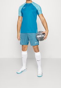 Hombre en ropa deportiva azul sosteniendo un balón de fútbol Nike F.C. negro y gris, con calcetas blancas y zapatos de interior Nike blancos.