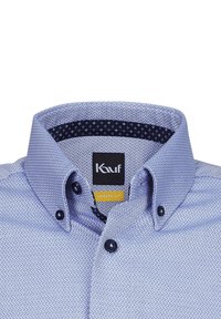 Camicia azzurra chiara con una trama texturizzata, caratterizzata da un colletto blu scuro con piccoli crocetti bianchi e bottoni neri.