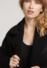 Vêtements d'extérieur noir, texturé, en matériau doux similaire à de la polaire et avec un large col. Le modèle porte un débardeur noir en dessous.