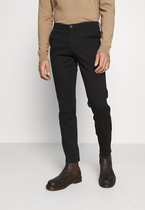 Mand iført slanke sorte bukser, brune Chelsea-støvler og en beige langærmet sweater, stående foran en ensfarvet hvid baggrund.