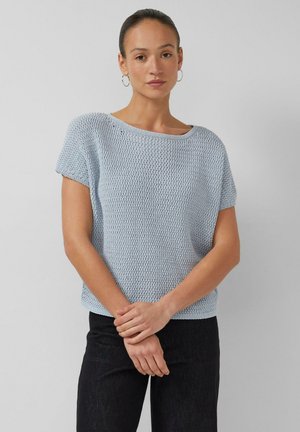 Kobieta w jasnoniebieskim teksturowanym swetrze z krótkimi rękawami i czarnych spodniach, stojąca z złożonymi rękami na jednolitym szarym tle.