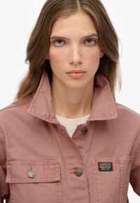 Jasnoróżowa kurtka z płótna z kołnierzem, dwoma kieszeniami na piersi i zatrzaskami. Zawiera tkane etykiety marki na lewej kieszeni.