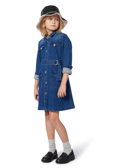 Jeune fille dans une robe en denim bleue ceinturée, portant des chausettes gris clair, des mocassins noirs et un chapeau seau noir et blanc, debout et regardant sur le côté.