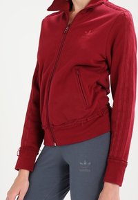 Červená sportovní bunda na zip vyrobená z jemné látky, s ribbovanými manžetami, dvěma bočními kapsami a třemi pruhy podél rukávů.