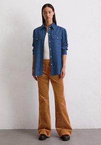 Donkerblauwe spijkerhemd met knopen en borstzakken, gecombineerd met oranje flared corduroy broek. Onder het hemd wordt een eenvoudig wit tanktop gedragen.