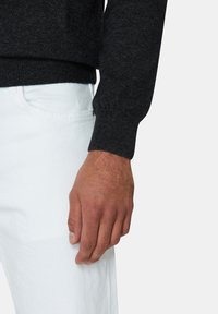 Dunkelgrauer Strickpullover mit gerippten Bündchen, kombiniert mit weißen Jeans aus Denim. Sichtbare Hand zeigt natürliche Hauttextur und Nageldetails.