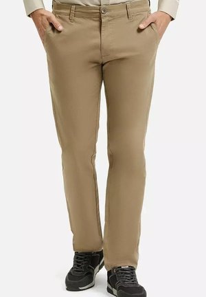 Beżowe bawełniane spodnie o prostym kroju, z kieszeniami z przodu i zapinane na guzik, w połączeniu z czarnymi trampkami. Gładka tekstura.