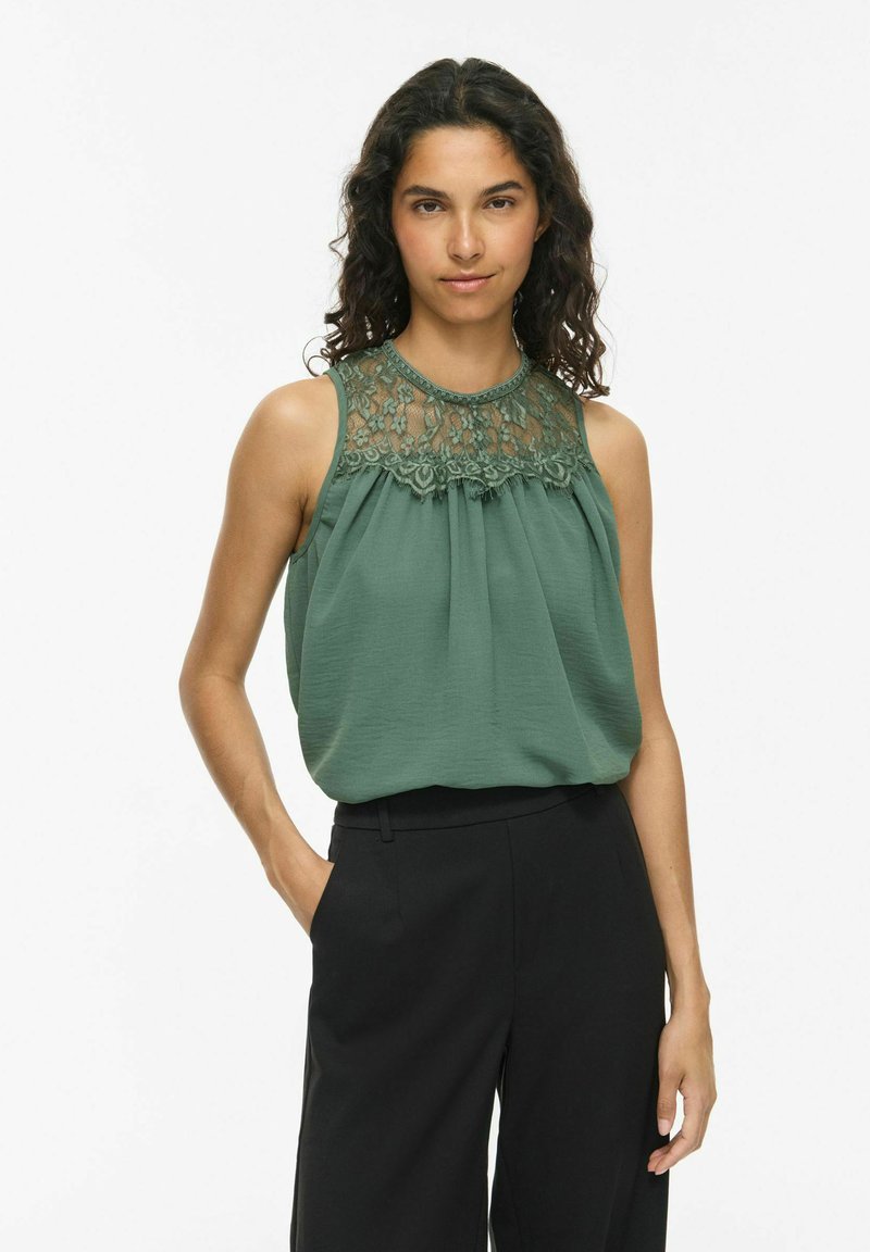 Femme aux cheveux bouclés et foncés portant un haut vert sans manches avec un détail en dentelle et un pantalon noir taille haute, debout avec une main dans la poche.