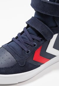 Zapatillas de deporte azul marino con material de ante y sintético, con acentos en rojo y blanco, cierre con cordones y Velcro, y diseño de punta perforada.