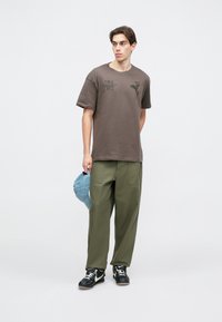 Καφέ βαμβακερό μπλουζάκι με γραφικό σχέδιο, συνδυασμένο με πράσινο παντελόνι σε φαρδιά γραμμή. Κρατάει ένα ανοιχτό μπλε καπέλο και φοράει μαύρα αθλητικά παπούτσια με λευκές λεπτομέρειες.