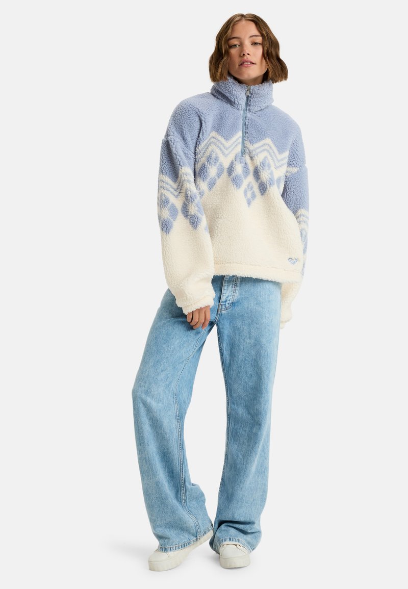 Pull en polaire bleu clair et crème avec un col zippé à mi-hauteur, des motifs géométriques et une coupe décontractée, associé à un pantalon en jean bleu clair à jambes larges.