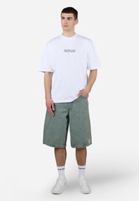 Fehér, oversized póló fekete hímzett szöveggel, világoskék farmer rövidnadrág, fehér sportcipő, fehér zokni fekete logóval, laza szabás.