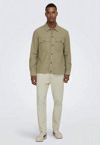 Camisa verde-oliva de botões com dois bolsos no peito, combinada com calças de cor creme e ténis bege, usada por uma figura masculina.