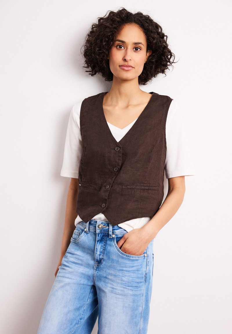 Frau mit lockigem Haar, die eine braune, geknöpfte Weste über einem weißen Hemd und hellblauen Jeans trägt, steht vor einem einfarbigen hellen Hintergrund.