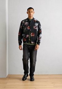 Μαύρη bomber jacket με λουλουδάτο σχέδιο, ελαιοπράσινη στρώση από κάτω, σκούρα τζιν και μαύρες μπότες. Το υποκείμενο στέκεται σε ξύλινο πάτωμα.
