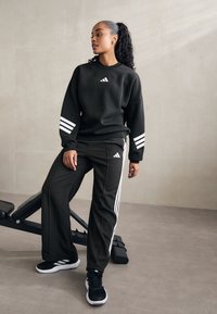 Sweatshirt noir et pantalon avec un design à trois rayures blanches. Tissu doux, coupe décontractée. Des chaussures de sport noires complètent la tenue.