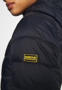 Marineblaue Steppjacke mit gestepptem Design und einem gelben Barbour International Logo-Patch am Ärmel. Glatte, glänzende Textur.